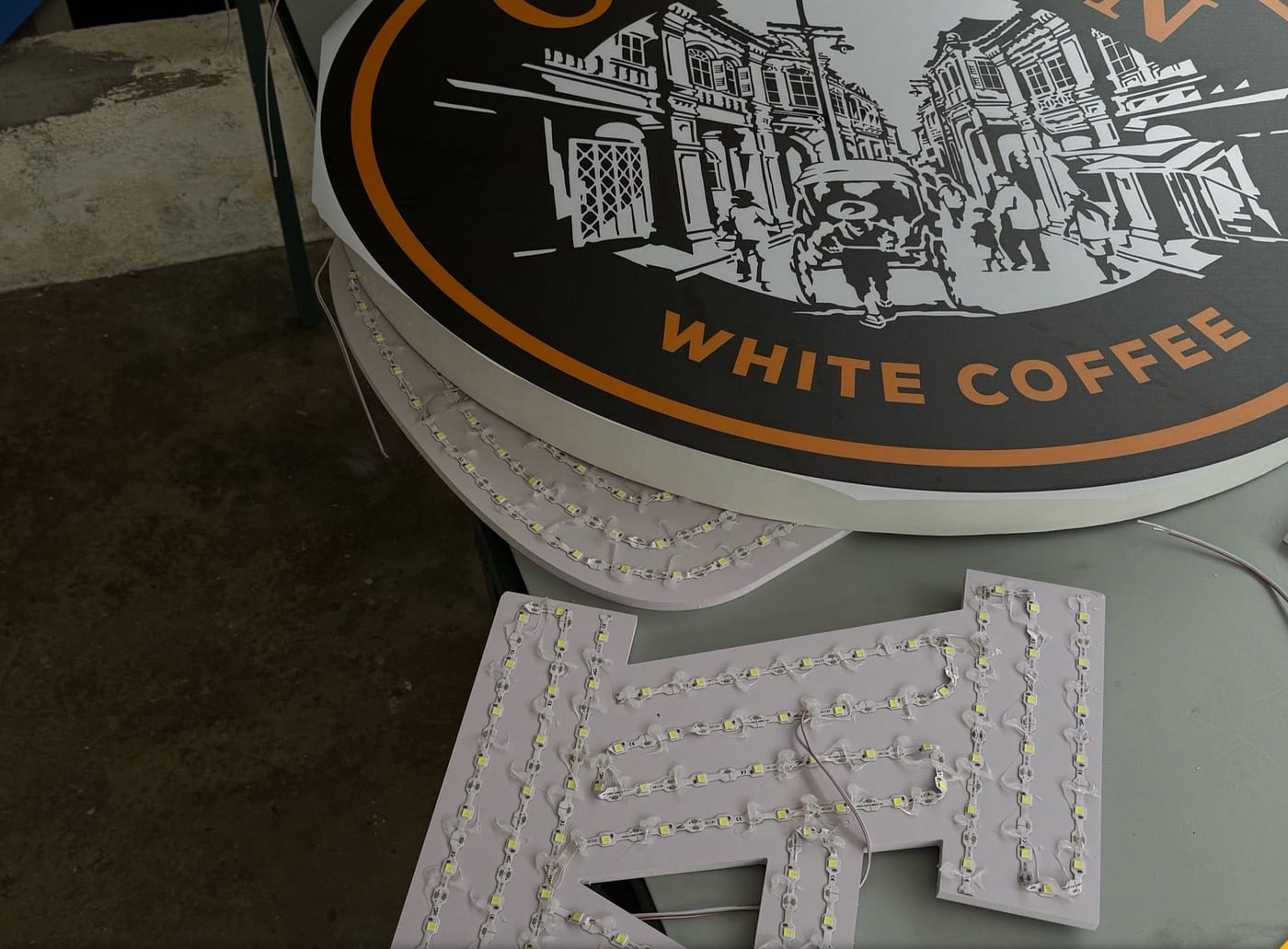 A completed circular OLDTOWN White Coffee signboard standing on trestles in a workshop, with loose white three-dimensional letters on the floor in front of it.