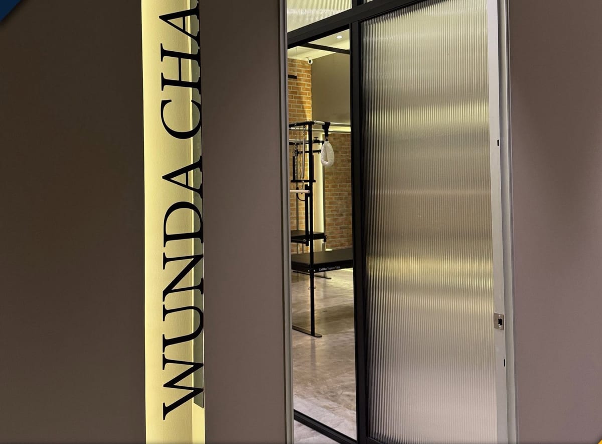 A slim vertical backlit panel reading WUNDA CHAIR mounted on a corridor wall beside a fluted-glass door.