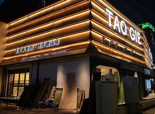 A corner building at night with a band of warm illuminated fins wrapping the upper facade and TAO GIE lettering on the fascia.